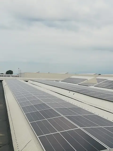 Falde di un capannone industriale coperte di moduli, con altri capannoni e le montagne