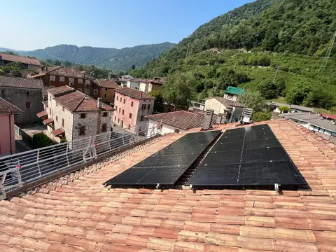 Due file di moduli neri su un tetto in coppi, con il borgo in pietra e le colline boscose intorno