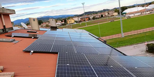 Moduli fotovoltaici su una copertura in lamiera rossa, accanto a un campo sportivo e alle colline