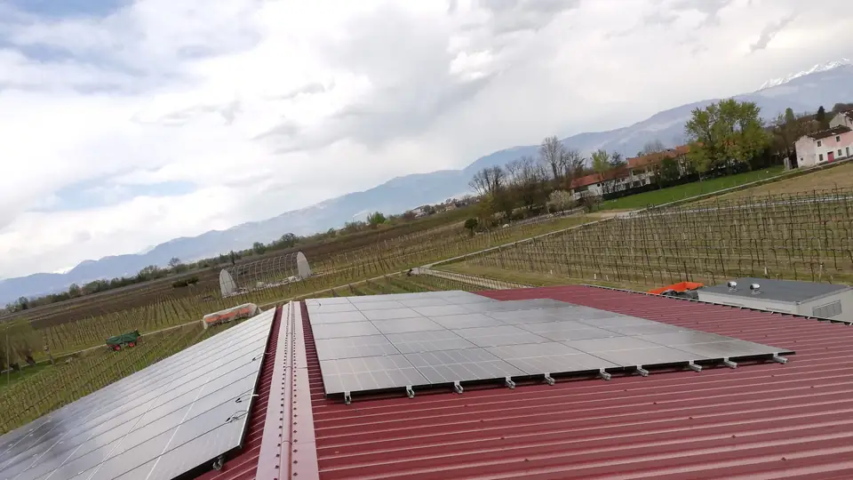 Moduli fotovoltaici su un tetto in lamiera rossa, tra i vigneti e le montagne